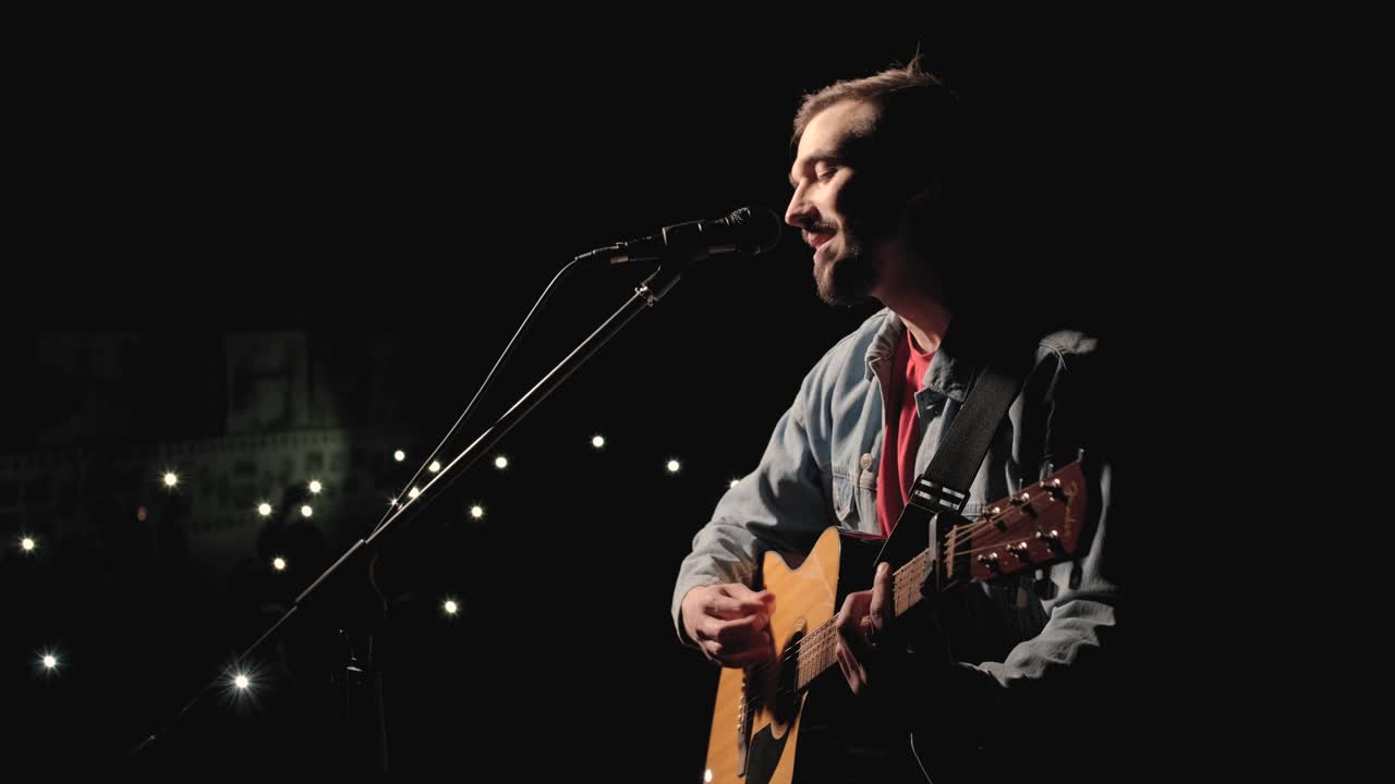 un músico está tocando la guitarra en un gran escenario con miembros de la audiencia sosteniendo linternas en el fondo. el auditorio está lleno de hermosas luces parpadeantes.