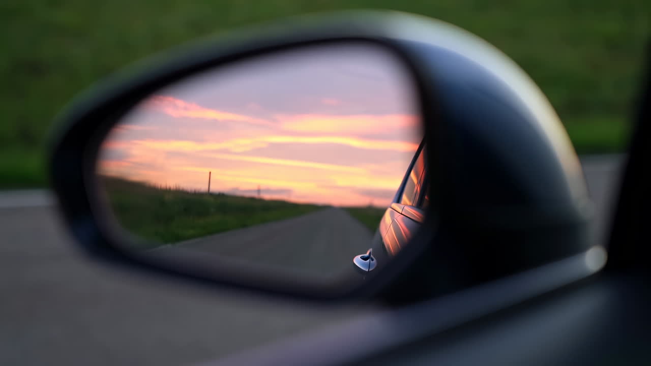 Sidespeil på bil som reflekterer solnedgangshimmel, med varme oransje og rosa fargetoner over et uskarpt landskap. Nærbilde.