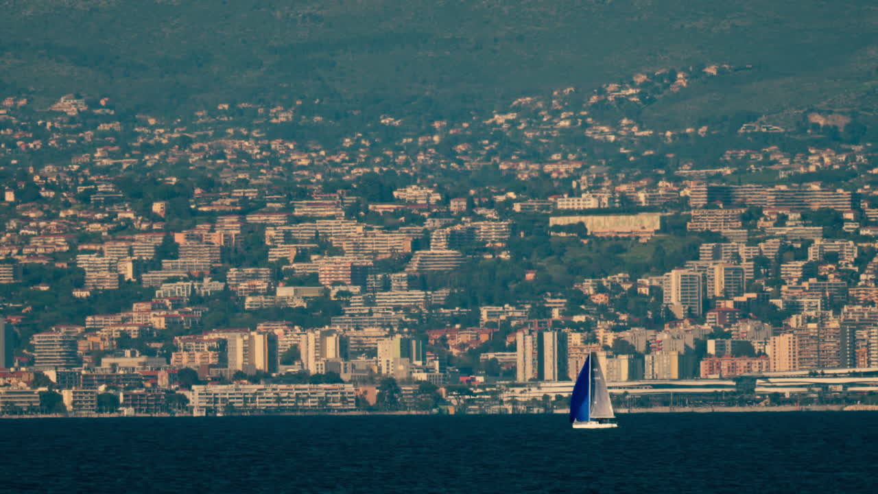 Distant view of a sail boat moving on the sea in Antibes, France with the city and the mountain on the background