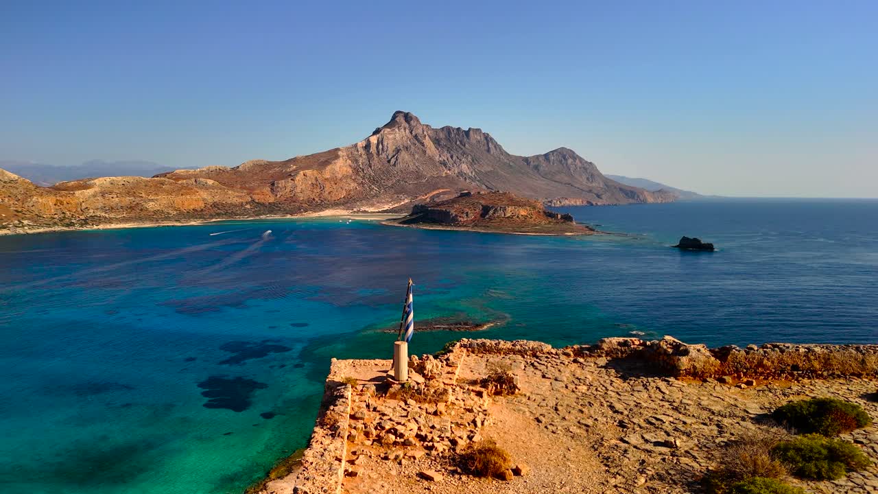 A breathtaking view of a Greek island's coastline and mountains taken from a high vantage point. The crystal blue waters sparkle under the midday sun, enhancing the natural beauty of the landscape
