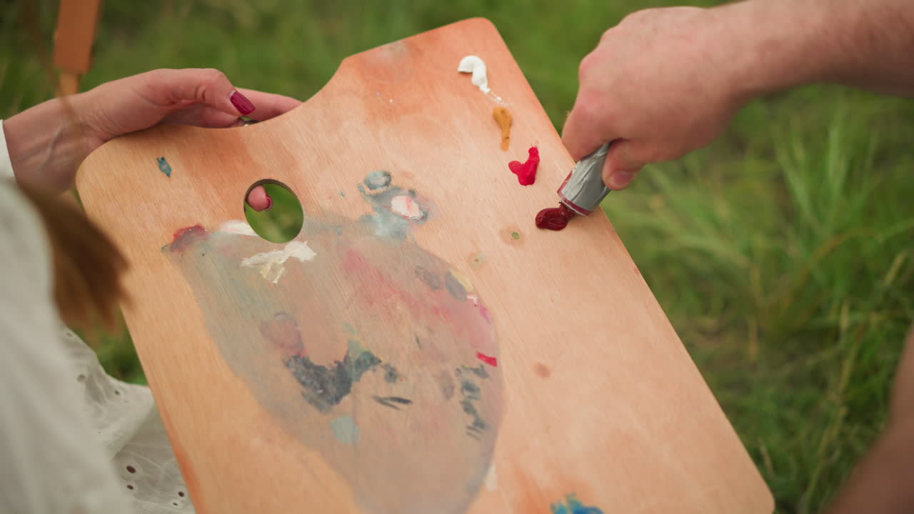 close-up of a collaborative artistic moment. A woman holds a wooden palette while a man carefully squeezes out paint from a tube onto the palette, preparing for a creative session