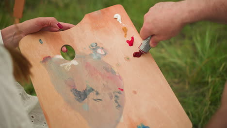 close-up of a collaborative artistic moment. a woman holds a wooden palette while a man carefully squeezes out paint from a tube onto the palette, preparing for a creative session