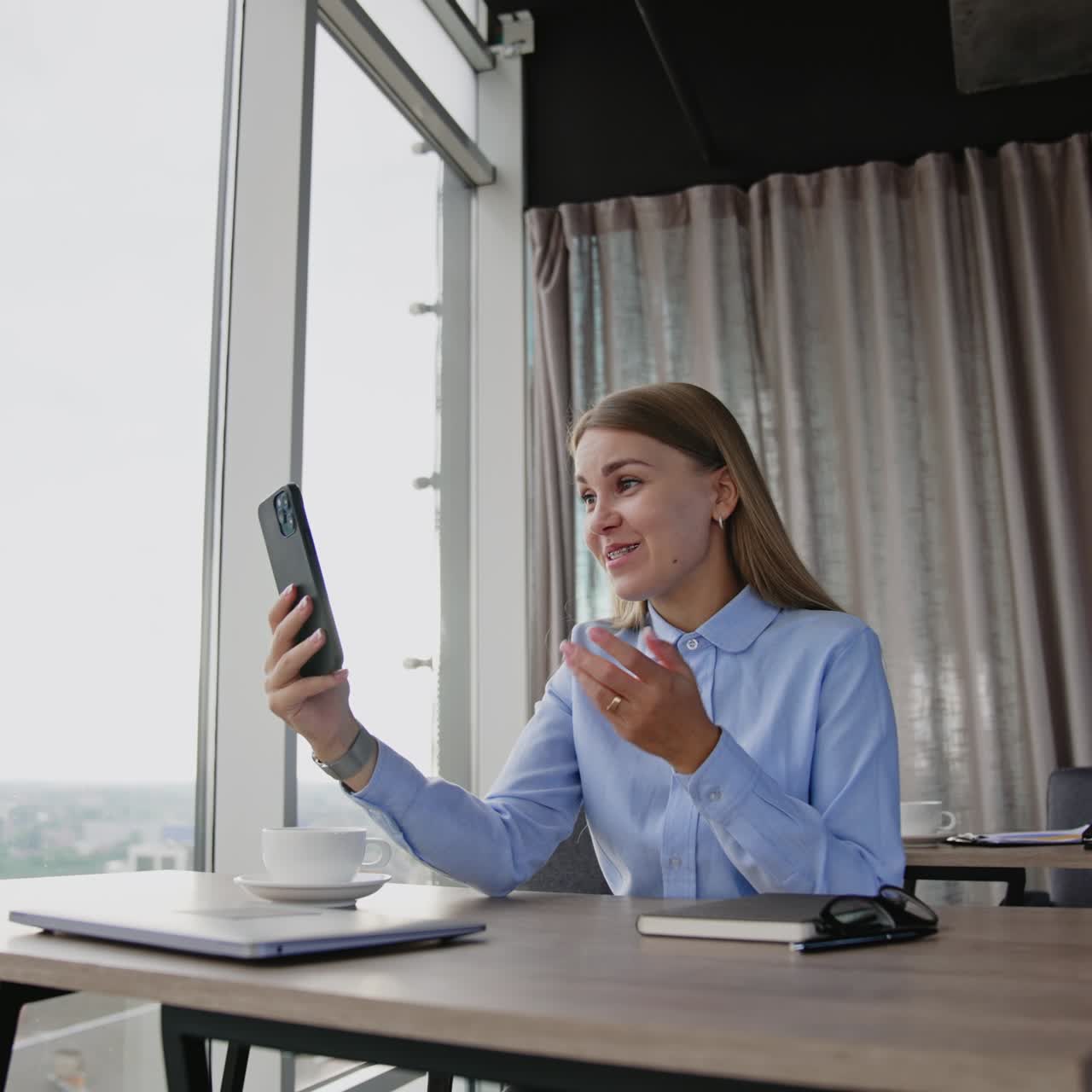Good-looking lady in blue shirt looking on the phone in front of her. Smiling woman having conversation via online video chat. Cityscape at backdrop in window