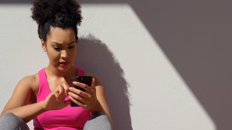 Front-view-of-young-mixed-race-woman-sitting-against-the-wall-in-backyard-of-home-4k