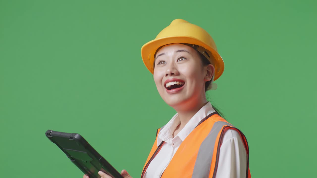 안전 헬을 착용한 아시아 여성 엔지니어가 손에 을 들고 녹색 스크린 배경 스튜디오에 서 있는 동안 주위를 둘러보고 있다.