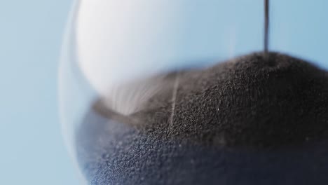 Video-of-close-up-of-hourglass-with-black-sand-pouring-with-copy-space-on-blue-background