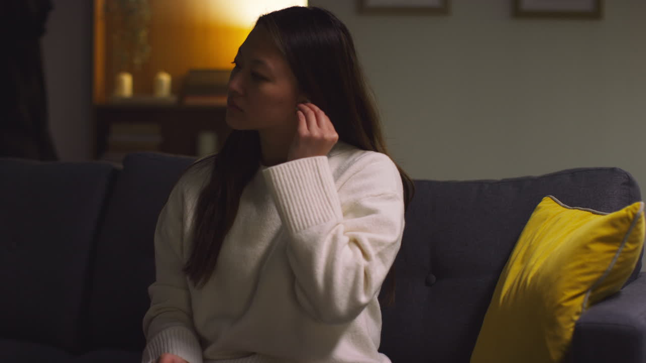 소파에서 집에서 저녁을 보내는 여성이 무선 이어폰을 착용하고 휴대 전화에서 음악이나 캐스트를 스트리밍합니다.