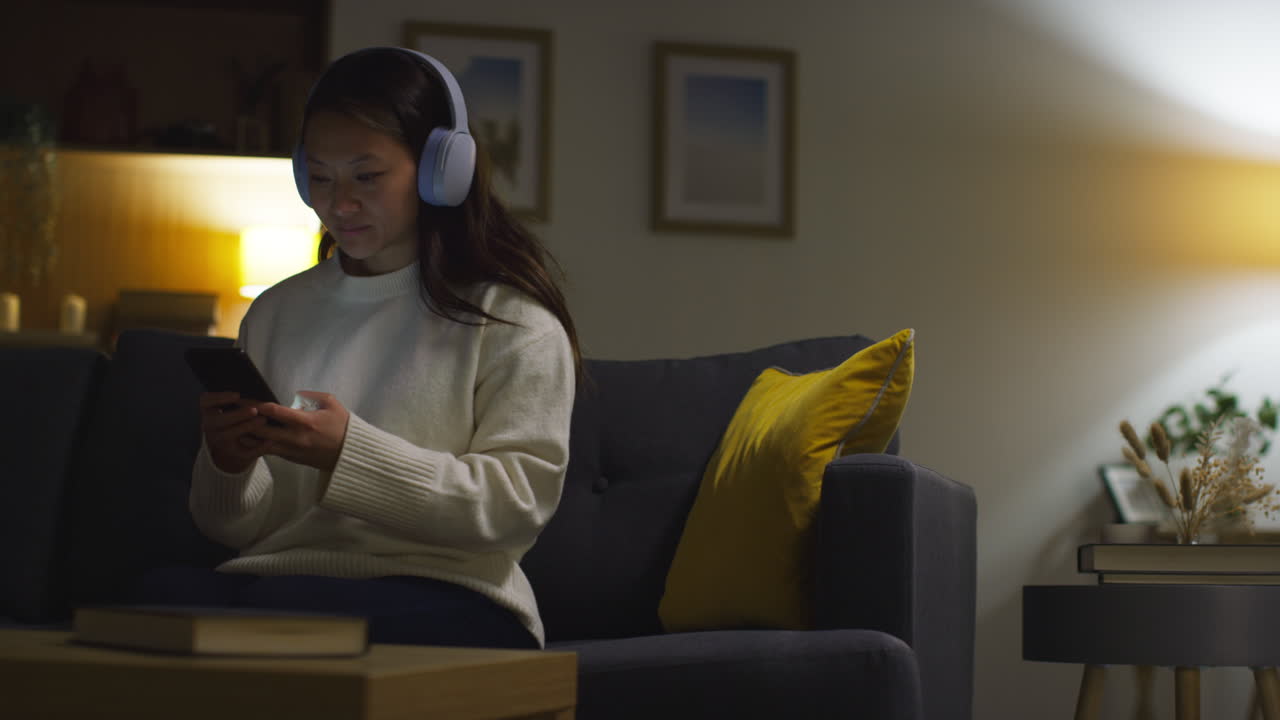 una mujer sentada en un sofá en una sala de estar, usando auriculares y mirando su teléfono