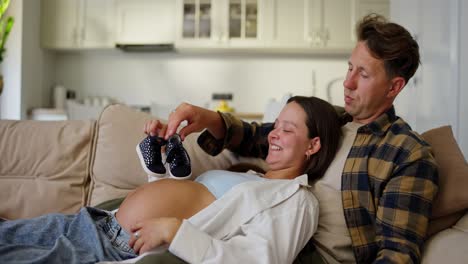 Glücklicher-Mann-In-Einem-Karierten-Hemd-Zusammen-Mit-Seiner-Schwangeren-Frau,-Die-Mit-Den-Kleinen-Schuhen-Eines-Neugeborenen-Babys-Zu-Hause-In-Einem-Modernen-Apartment-Spielt