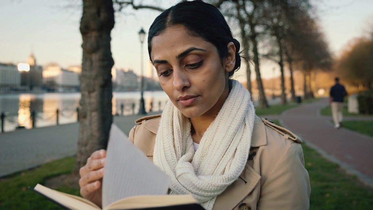 Reading woman in trench coat touching page, turning book on bench at riverside, locating passage