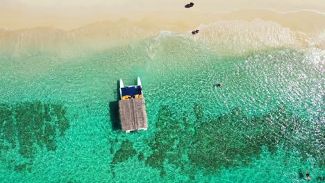 Touring-boat-with-straw-roof-floating-over-calm-clear-water-of-turquoise-sea-over-beautiful-pattern-of-rocky-seabed-and-coral-reefs-near-white-sandy-beach