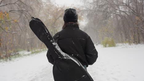 back view of person walking through snowy park carrying guitar pack on shoulder, surrounded by frosted trees and winter scenery, with benches and a misty path creating a tranquil atmosphere