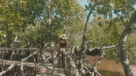 General-shot-of-a-European-young-woman-wearing-a-straw-hat-standing-alone-on-a-wooden-bridge-in-a-forest-enjoying-the-green-landscape