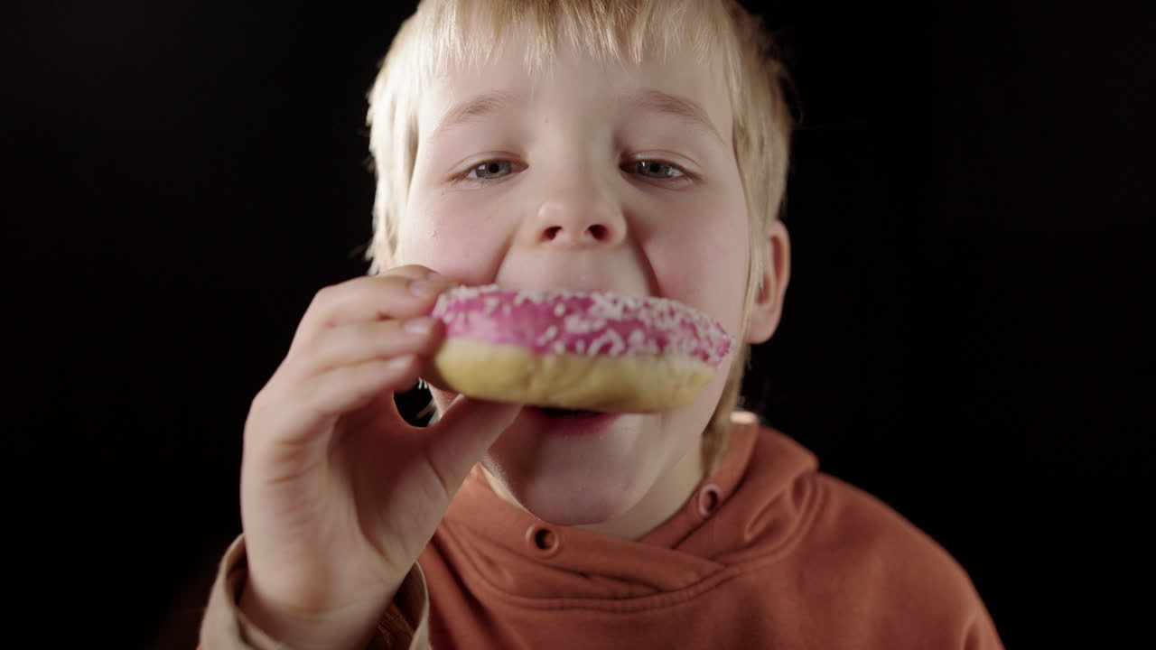 Close-up frontal of European boy biting into moist pink glazed donut