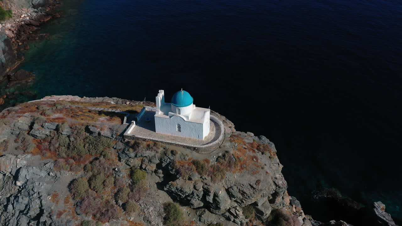 Aerial shot of a Greek orthodox blue dome chapel built on a cliff overlooking the Aegean sea. Camera tilts down while approaching the chapel from behind.