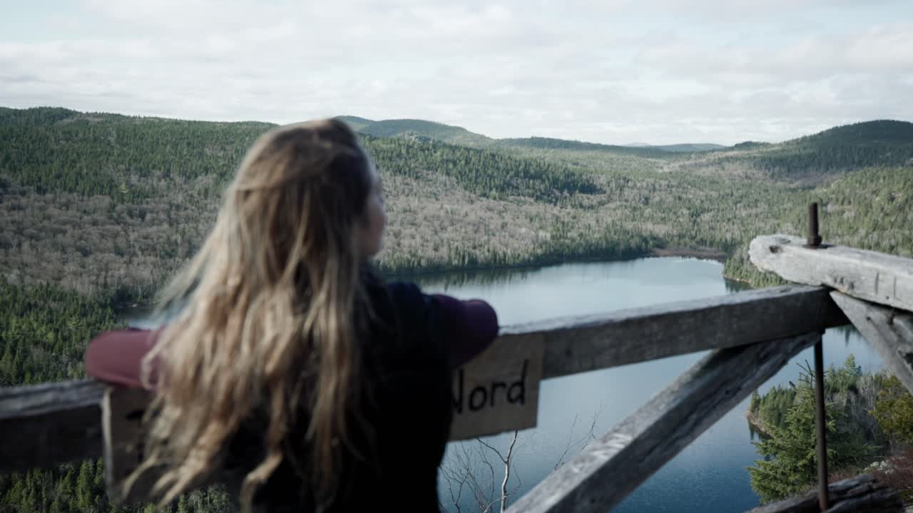 vista trasera de una joven rubia parada en una plataforma de observación de madera, disfrutando de la vista sobre el lago del bosque en saint-come, quebec, canadá