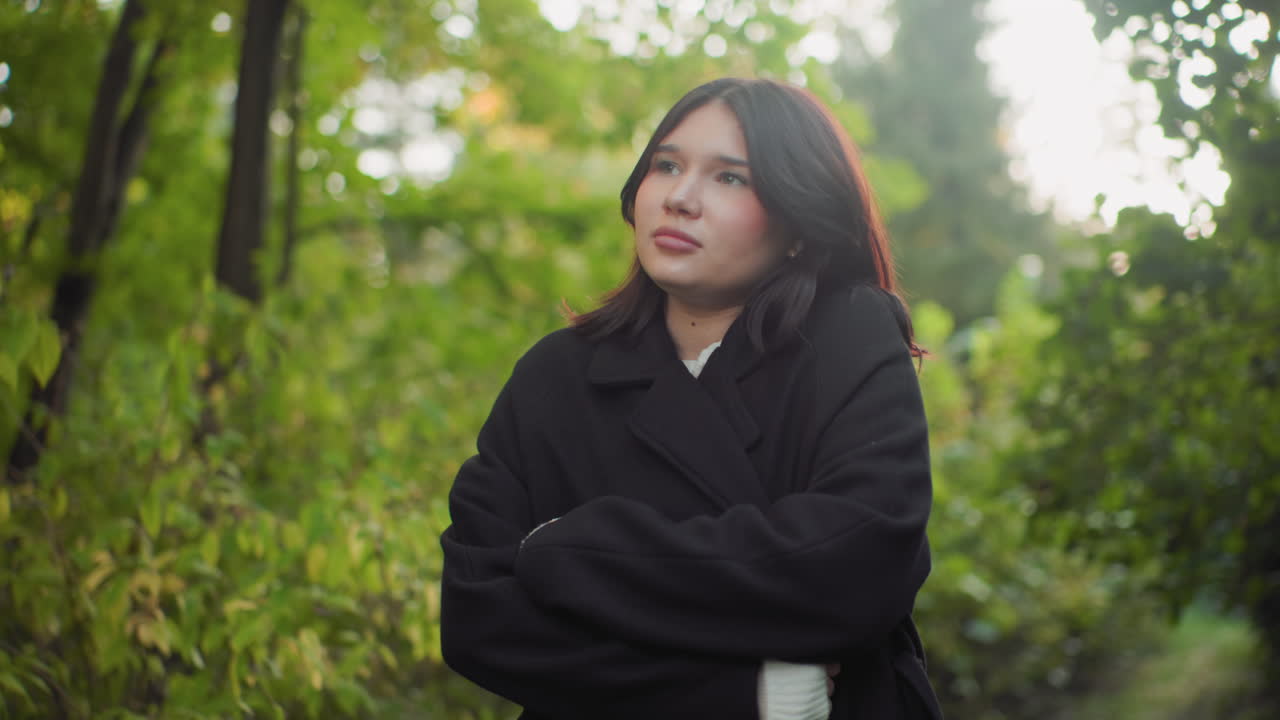 Shivering girl adjusting coat while walking through green forest path, looking around with calm pensive gaze, cheeks flushed from cold, soft daylight on leaves, gentle breeze across jacket sleeves