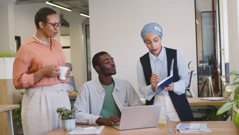 Junger-Arbeiter,-Der-Mit-Einem-Laptop-Arbeitet,-Der-An-Seinem-Schreibtisch-Sitzt,-Während-Muslimische-Geschäftsfrau-Und-Geschäftsfrau-Mit-Ihm-Im-Stehen-Sprechen