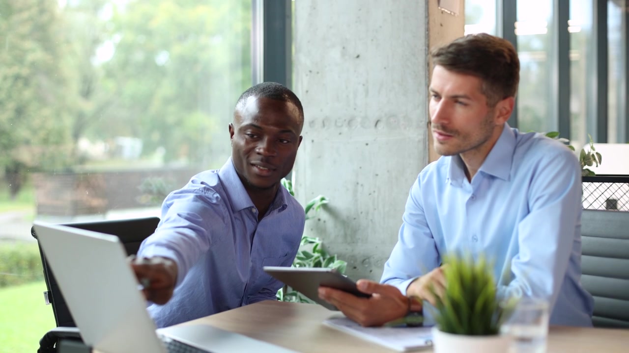 Two Diverse Businessmen Collaborating on a Tablet and Laptop in a Modern Office