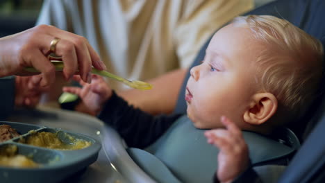 feeding a baby from spoon. female hand shoves the spoon to a cute little blond child. close up side view.
