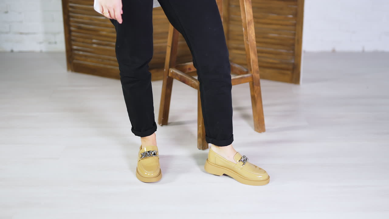 Woman in black jeans stands up from a wooden stool to demonstrate new shoes. Beige fashionable loafers with chain decoration presentation.