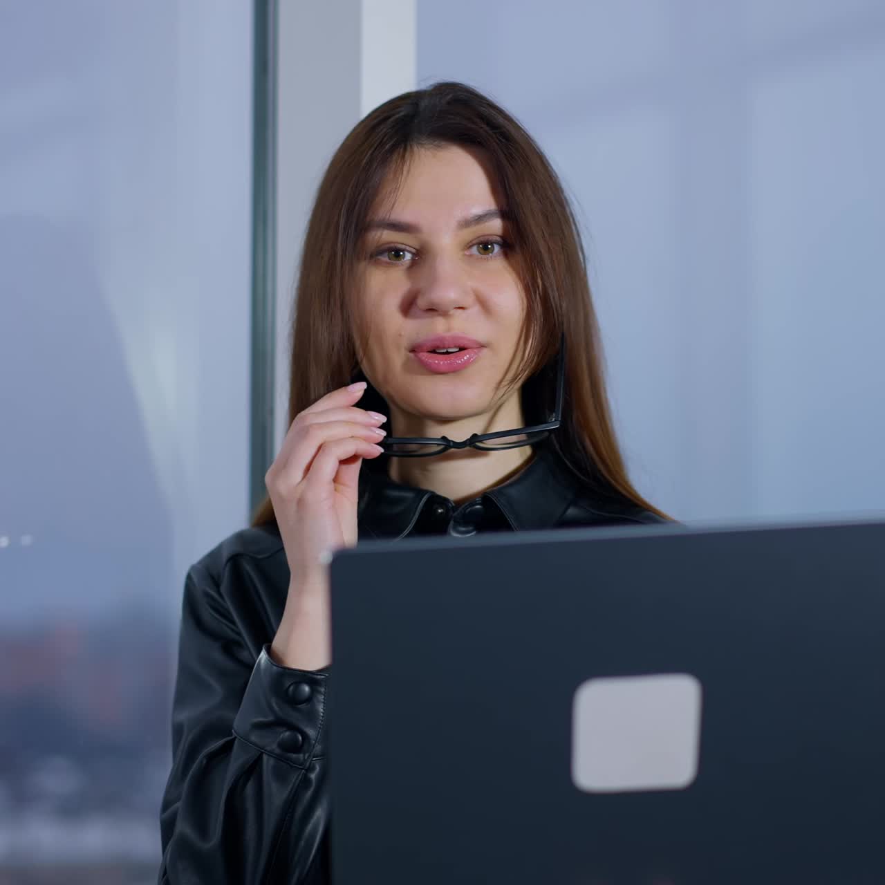 Busy female entrepreneur puts on glasses looking at laptop in her hands. Lady stands at the window ready to start online skype meeting