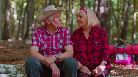 grandmother grandfather senior tourists hikers sitting on tree and hugging, kissing in summer wood