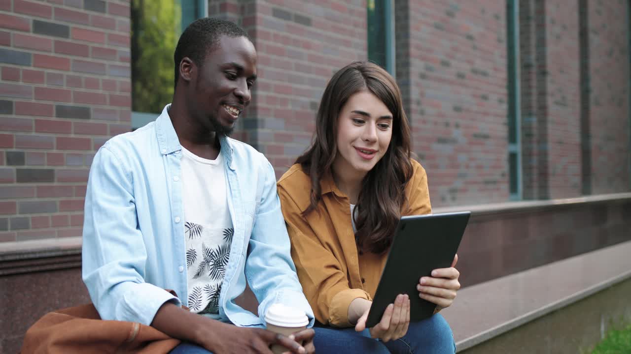 hombre afroamericano y mujer caucásica sentados en la calle y haciendo una videollamada en una tableta cerca de la universidad
