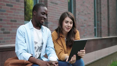 african american man and caucasian woman sitting in the street and having a video call on tablet near the college