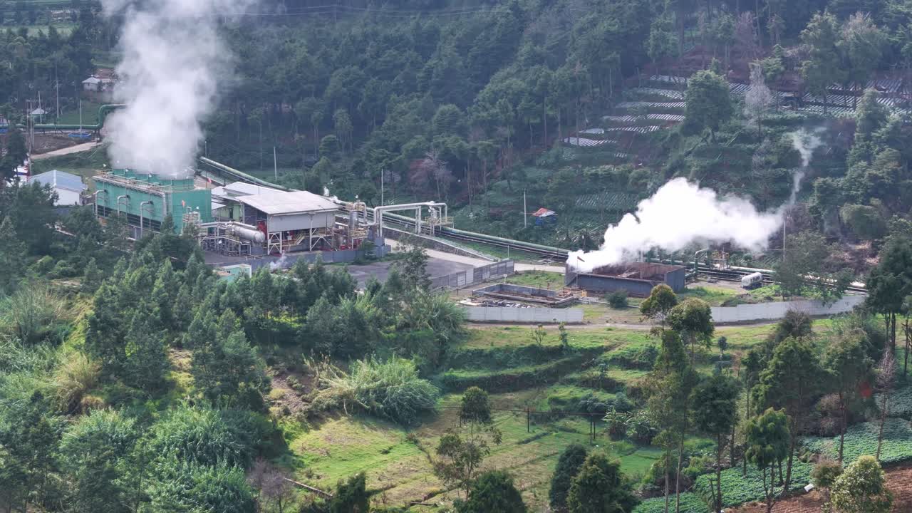 Aerial footage of a geothermal power plant surrounded by green hills, with steam rising from industrial facilities generating renewable energy