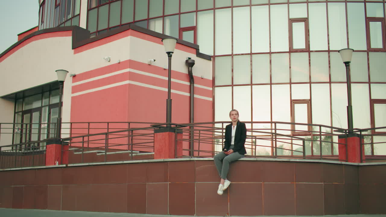 Young woman in formal outfit seated on fence with legs crossed at ankle, looking at phone in hands, surrounded by modern glass building and lamp posts in urban environment
