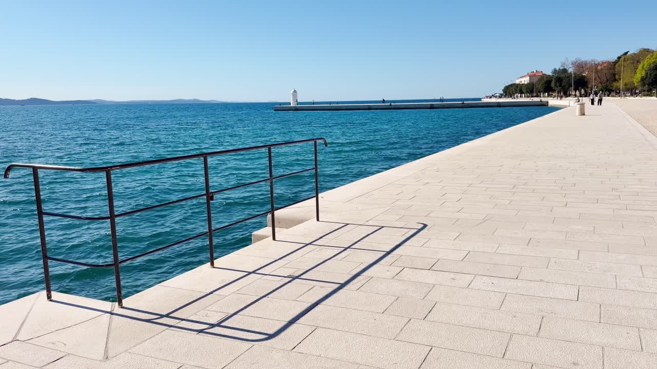 Pemandangan tenang dan cerah perlahan di promenade batu putih Zadar dengan orang-orang yang tidak dapat dikenali di kejauhan, mercusuar putih kecil, air Adriatik yang berkilauan, dan pegangan tangan hitam yang membentuk bayangan.