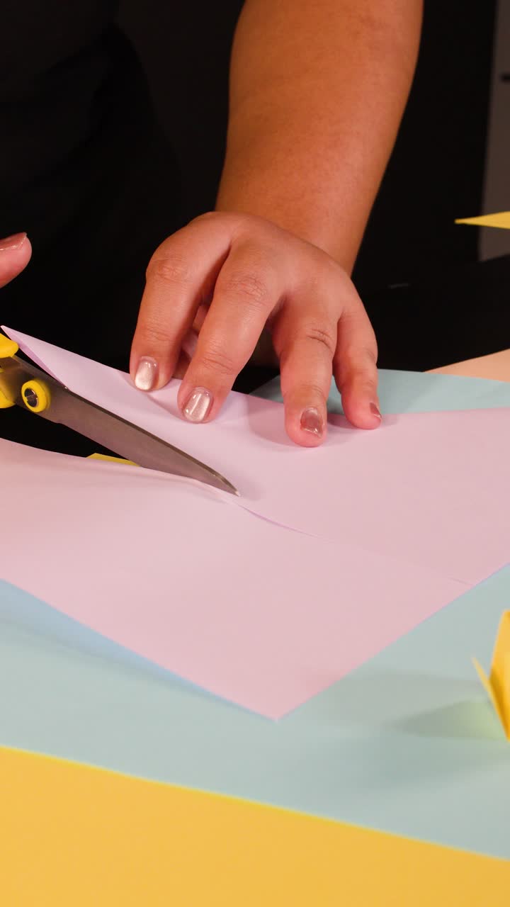A person uses yellow scissors to carefully cut pink paper on a colorful craft table