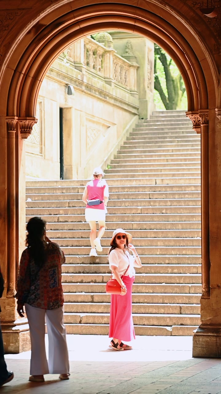 New York, USA, 22 maggio 2026: Donna in posa sotto l'arco di Bethesda Terrace, Central Park, New York. Arco in arenaria dai toni caldi che incornicia una figura in gonna rosa su gradini illuminati dal sole.