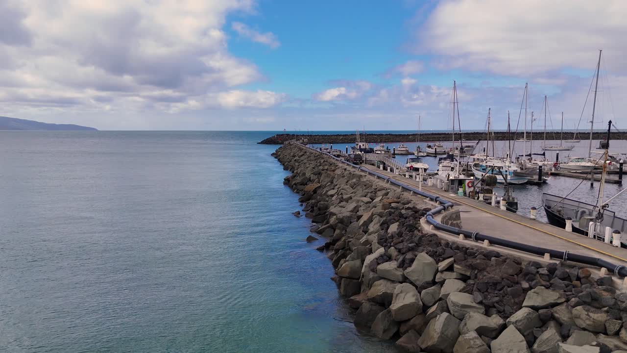 Drone footage captures Apollo Bay's harbor, showcasing boats, rock walls, and a jetty under a partly cloudy sky