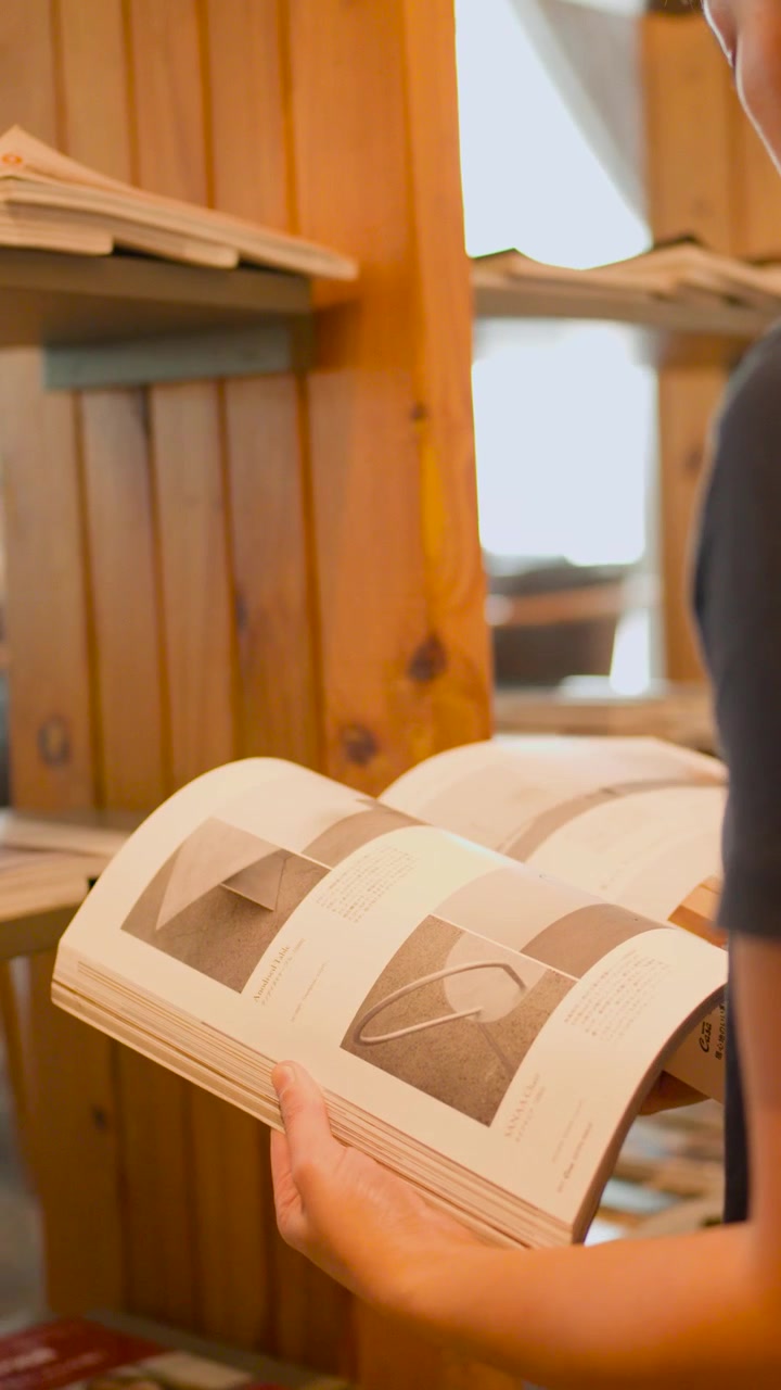 Una persona sfoglia una rivista mentre è in piedi accanto a una libreria di legno in una luce naturale soffusa.