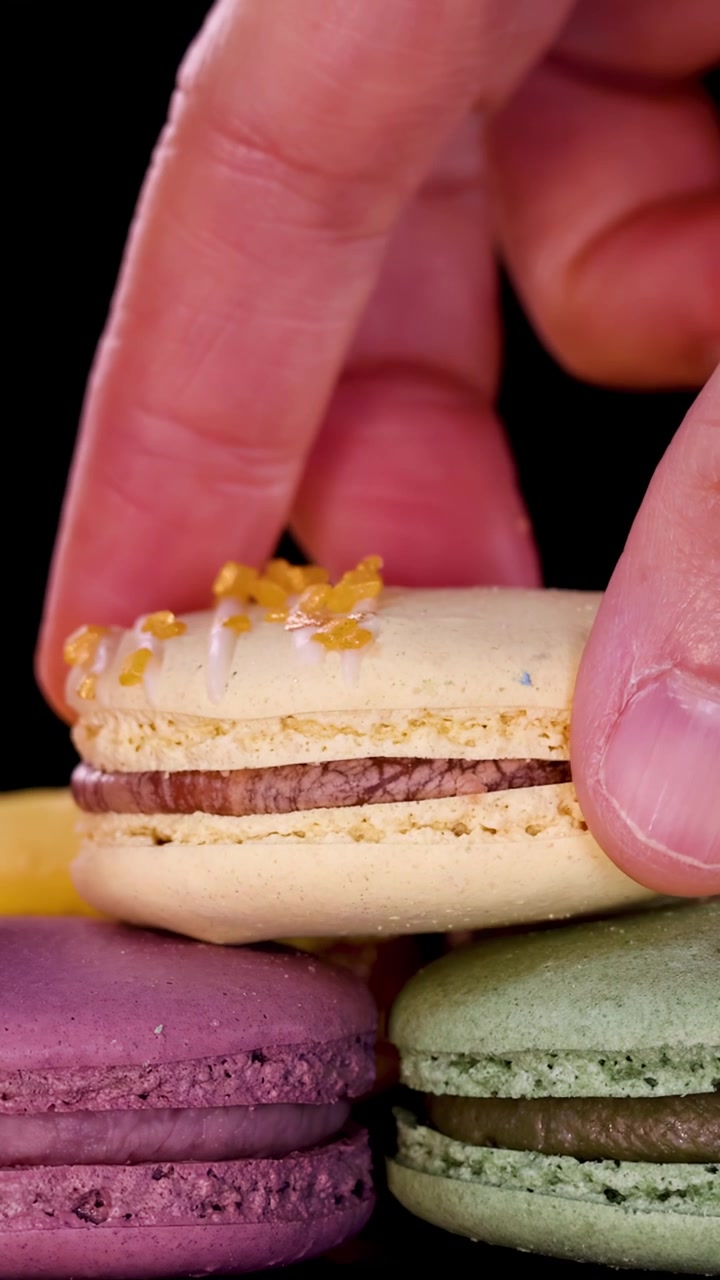 A close-up shot of a hand picking up a yellow macaron from a neat row of assorted colorful macarons on a dark background, with soft studio lighting and smooth camera movement