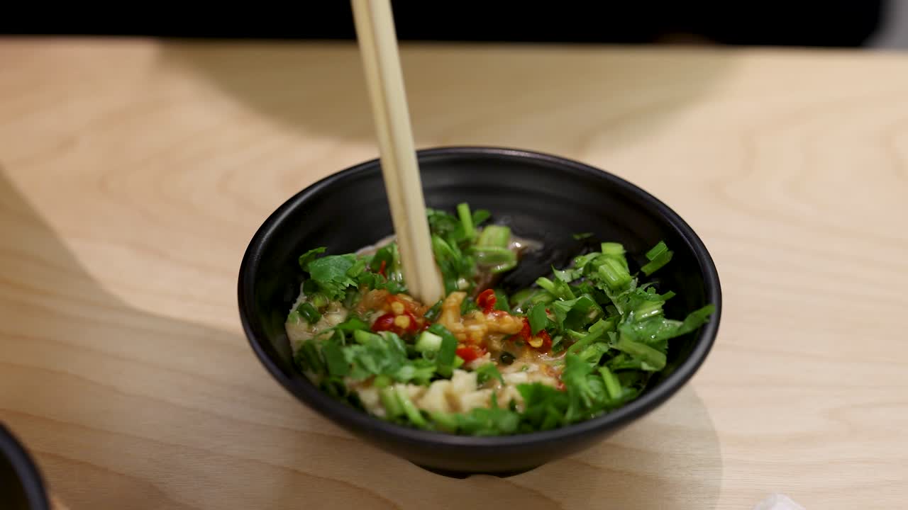 Hand stirs vibrant Asian chili garlic sauce with herbs in black bowl under soft lighting