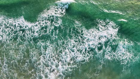 overhead drone footage captures dynamic, foamy ocean waves crashing onto a sandy shoreline in daylight, with vibrant turquoise water and textured surf patterns