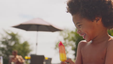 Grandfather-And-Grandson-Eating-Ice-Lollies-At-Edge-Of-Swimming-Pool-On-Family-Summer-Holiday