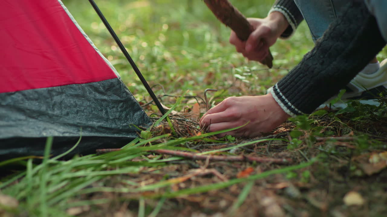 close up partial view of camper crouched on forest floor using short wooden stick to drive metal peg into ground beside red tent surrounded by grass leaves and pine needles during camp setup