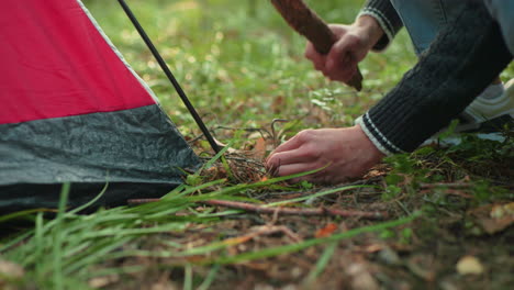 close up partial view of camper crouched on forest floor using short wooden stick to drive metal peg into ground beside red tent surrounded by grass leaves and pine needles during camp setup