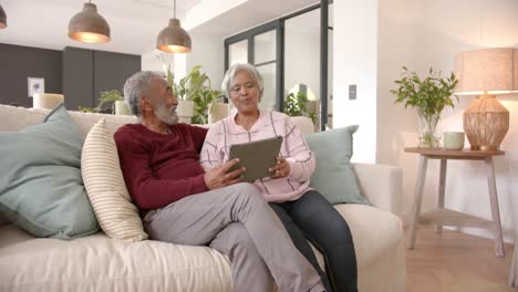 happy senior biracial couple sitting on sofa and using tablet, unaltered, copy space, in slow motion
