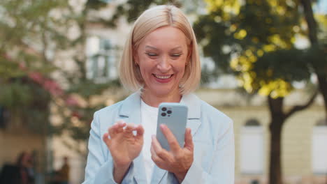 mature caucasian businesswoman ceo celebrating good news on smartphone standing on city street