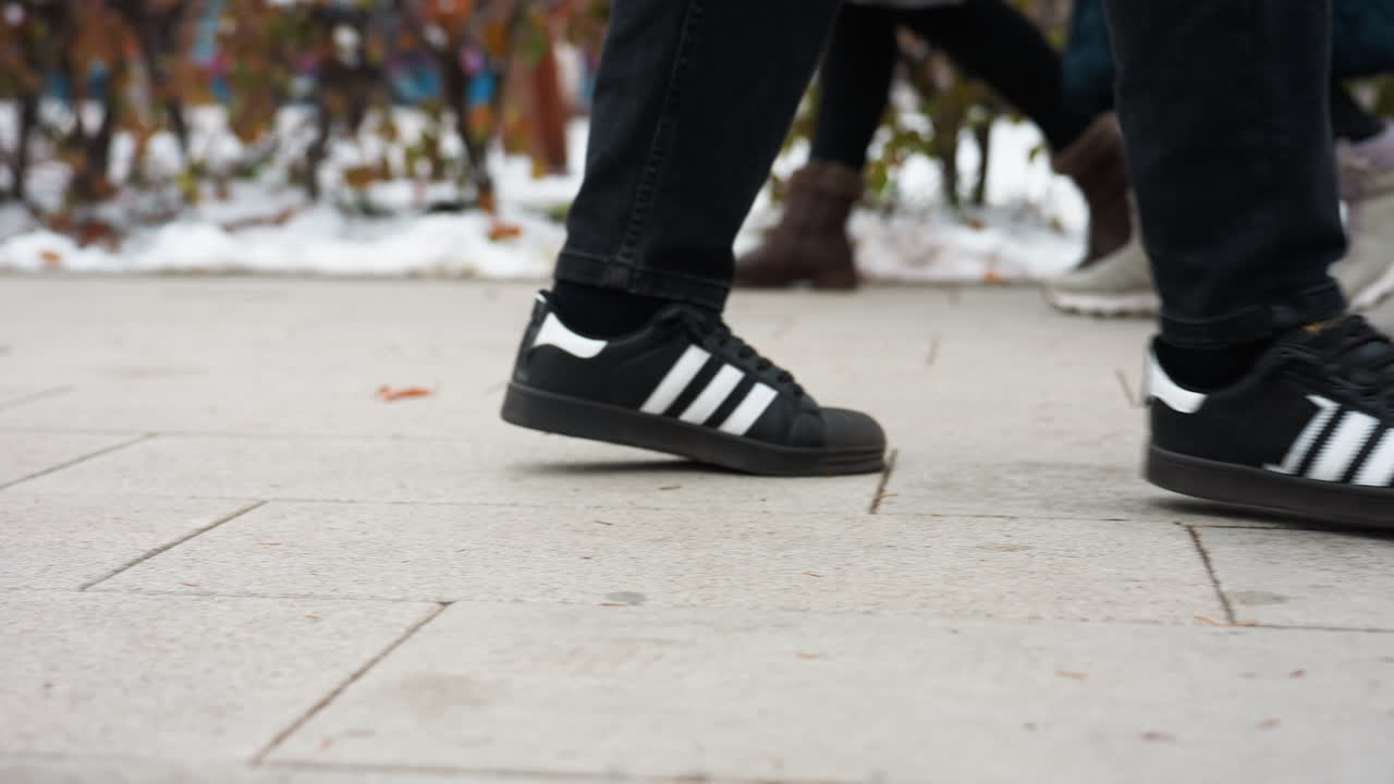 vista lateral de la pierna de un individuo con pantalones negros y zapatillas de deporte con rayas blancas caminando por un camino entrelazado el fondo borroso presenta nieve, árboles y una figura poco clara con otras personas que pasan