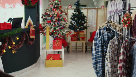 dolly in shot of festive clothing store filled with beautifully decorated christmas tree, tinsel garlands, red package presents and other ornaments during winter holiday season