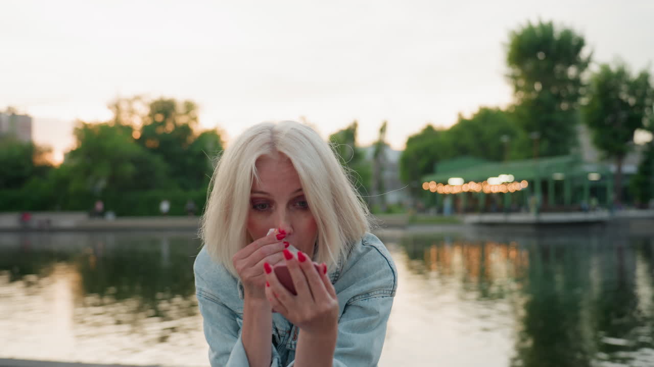 elegant lady seated on bridge by river park uses handheld mirror and facial wipe to remove bright pink paint from nose after playful picnic painting session at sunset, serene water background