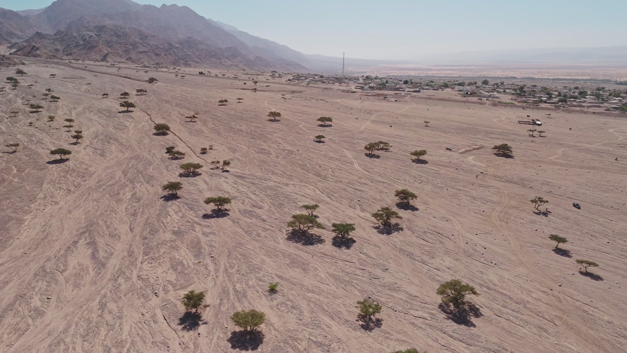 Side drone of red rocky desert with trees and village and mountains in valley