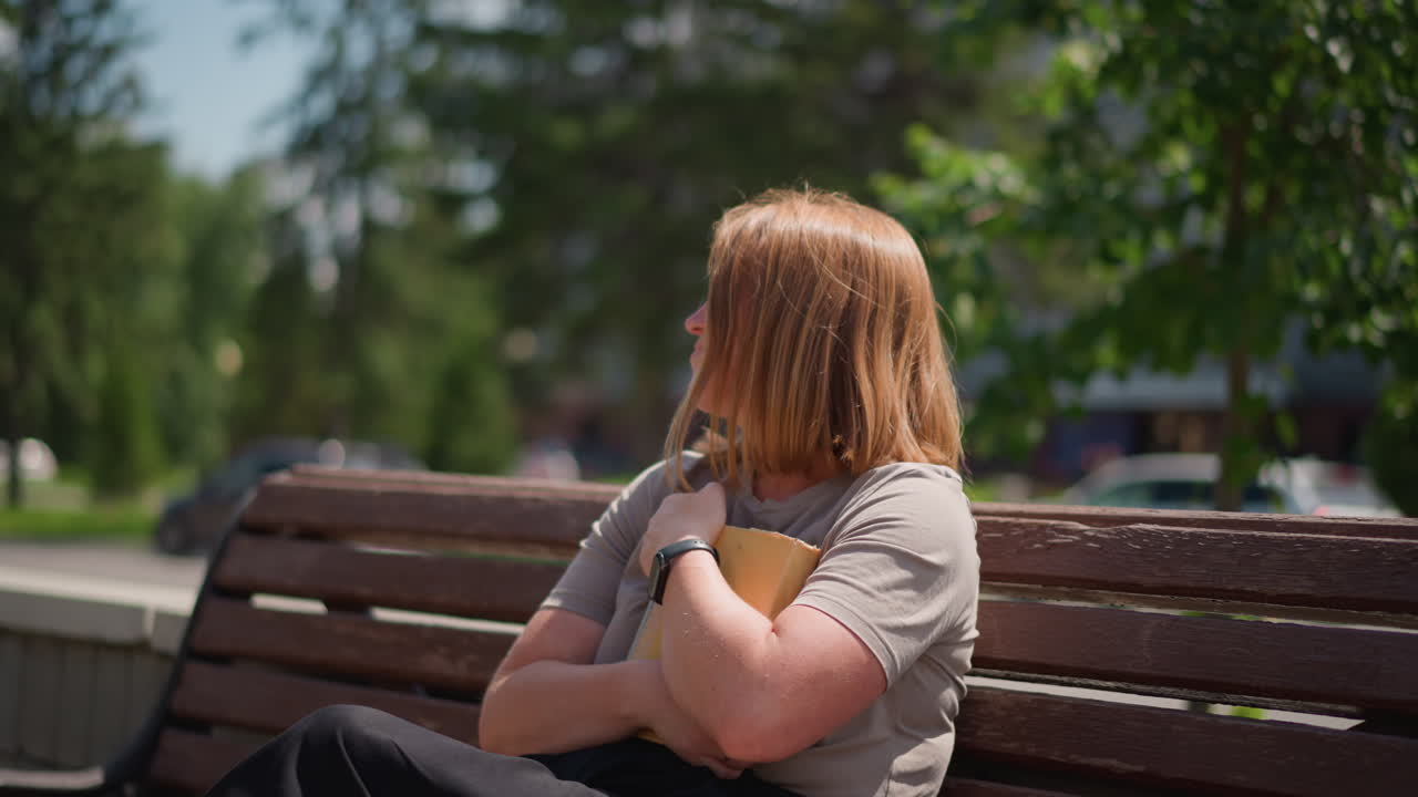 Student sits on wooden bench reading under sunlight in urban park then closes book and presses it to chest while pigeon flies and car passes nearby calm reflective summer moment with greenery