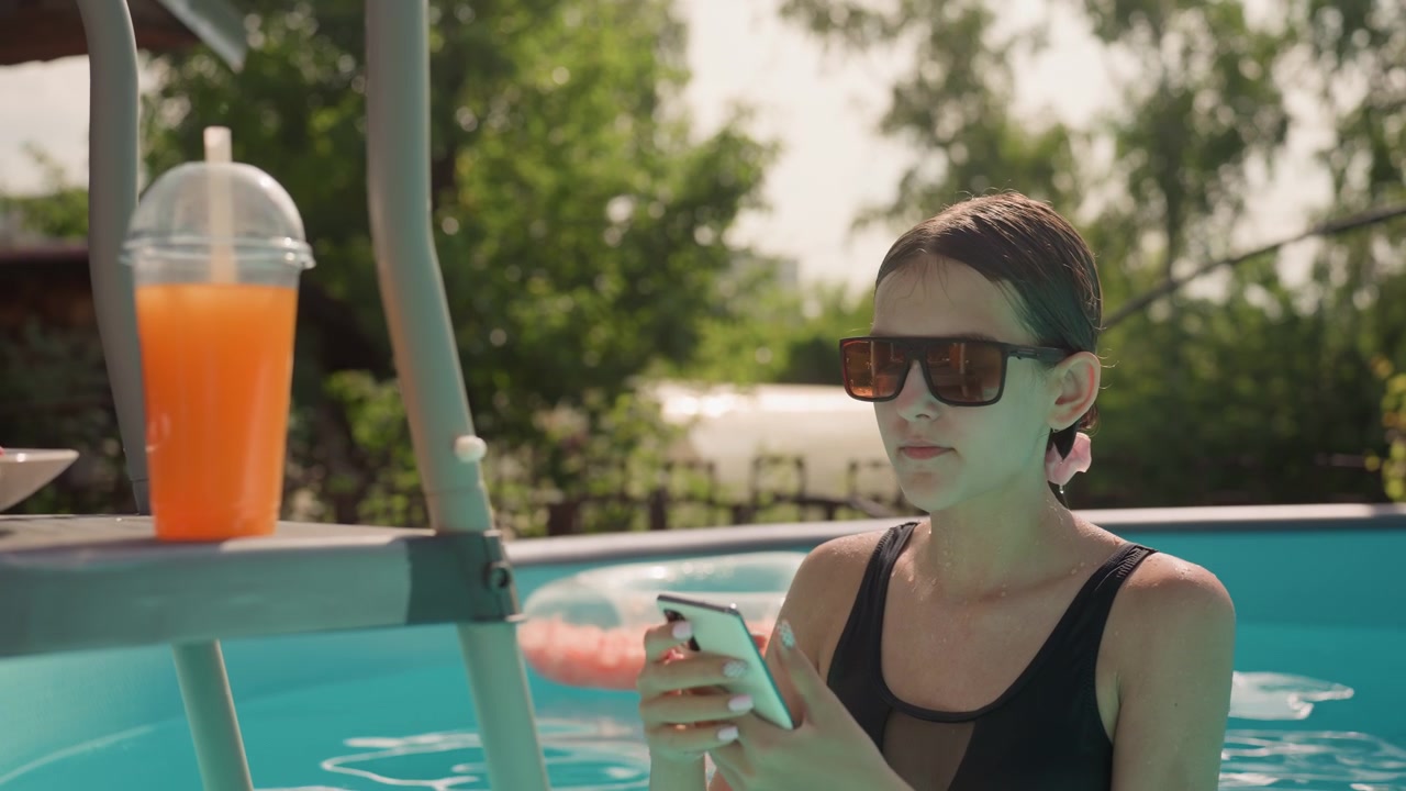Woman in black swimsuit relaxes by outdoor pool reaching for her phone on ladder while holding onto it. orange drink and bowl of cherries sit nearby as woman interacts with both objects